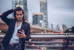 Image of a man with long hair wearing a suit and using a smart phone on a bridge in front of a cityscape